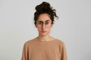 retrato de grave concentrado joven mujer con bollo de oscuro Rizado pelo usa beige camisa de entrenamiento y lentes mira estricto un confidente aislado terminado blanco antecedentes foto