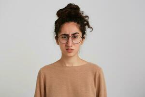 Closeup of confused unhappy young woman with bun of dark curly hair wears beige sweatshirt and glasses feels displeased and disappointed isolated over white wall photo