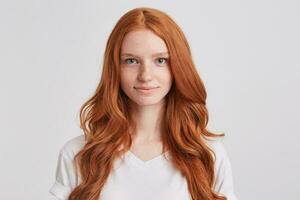 Portrait of smiling attractive redhead young woman with long wavy hair and freckles wears t shirt feels happy and satisfied isolated over white background Looks directly in camera photo