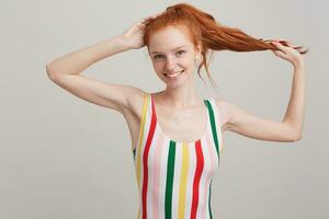 Portrait of happy beautiful young woman with red hair and freckles in striped top holding her ponytail and posing isolated over white background Looks satisfied photo