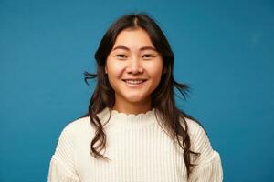 Glad attractive young brunette lady with romantic hairstyle smiling cheerfully while looking happily at camera, posing over blue background in knitted clothes photo