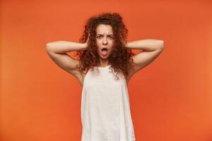 Portrait of attractive, adult redhead girl with curly hair. Wearing white off-shoulder blouse. Covering her ears with hands. Shock on a face. Watching at the camera, isolated over orange background photo