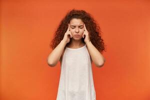 Nice looking woman, beautiful girl with curly ginger hair. Wearing white off-shoulder blouse. Keeps her eyes closed and massaging temples, headache. Stand isolated over orange background photo