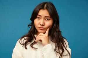 Pensive young pretty long haired asian female with natural makeup keeping index finger on her cheek while looking thoughtfully at camera, posing over blue background photo