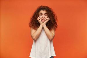 Teenage girl, terrifying looking woman with curly ginger hair. Wearing white off-shoulder blouse. Touching her face. Has fear on her face. Watching at the camera, isolated over orange background photo