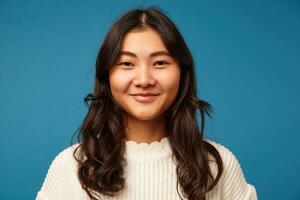 Horizontal shot of young charming dark haired asian lady with wavy hairstyle being in nice mood and smiling positively while standing over blue background photo