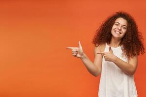 Portrait of attractive, adult redhead girl with curly hair. Wearing white off-shoulder blouse. Watching at the camera and pointing to the left at copy space, isolated over orange background photo