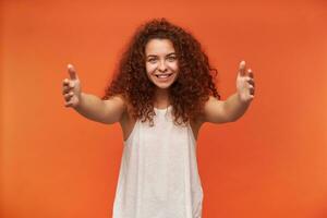 Portrait of attractive, lovely redhead girl with curly hair. Wearing white off-shoulder blouse. Spread her arms, trying to hug you. Watching at the camera, isolated over orange background photo