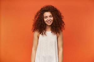 Portrait of attractive, adult redhead girl with curly hair. Wearing white off-shoulder blouse. Have messy hair and smiling. Watching at the camera, isolated over orange background photo