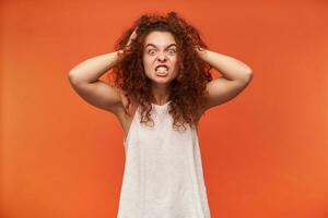 Angry looking woman, crazy girl with curly ginger hair. Wearing white off-shoulder blouse. Touching her head, terrible headache. Watching at the camera, isolated over orange background photo