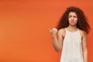 Portrait of unhappy, displeased redhead girl with curly hair. Wearing white off-shoulder blouse. Pointing with a thumb and watching to the left at copy space, isolated over orange background photo