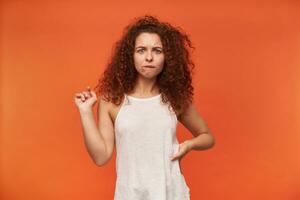 Nice looking woman, beautiful girl with curly ginger hair. Wearing white off-shoulder blouse. Holds hand on a waist and biting a lip. Watching bewildered at the camera, isolated over orange background photo