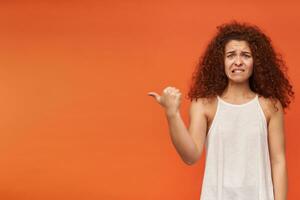 Nice looking woman, unhappy girl with curly ginger hair. Wearing white off-shoulder blouse. Biting a lip. Watching at the camera and pointing to the left at copy space, isolated over orange background photo