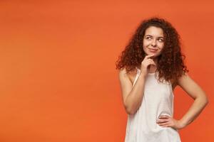 Portrait of attractive, adult redhead girl with curly hair. Wearing white off-shoulder blouse. Touching her chin and smile, wonder. Watching to the left at copy space, isolated over orange background photo