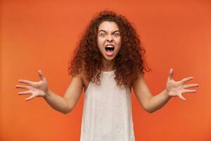 Portrait of evil, adult redhead girl with curly hair. Wearing white off-shoulder blouse. Trying to scare you. Screams and spreading her arms. Watching at the camera, isolated over orange background photo