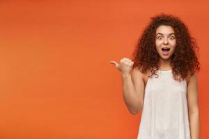 Surprised looking woman, cute girl with curly ginger hair. Wearing white off-shoulder blouse. Watching at the camera, pointing with a thumb to the left at copy space, isolated over orange background photo