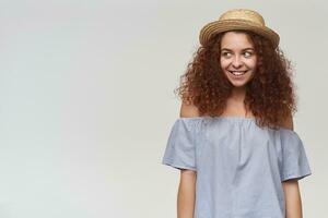 Portrait of attractive, adult redhead girl with curly hair. Wearing striped off-shoulders blouse and hat. Flirty smiling. Watching to the left at copy space, isolated over white background photo