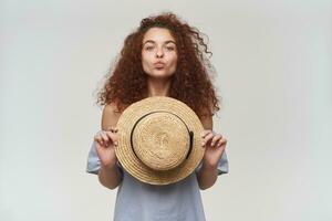 Portrait of attractive, adult redhead girl with curly hair. Wearing striped off-shoulders blouse and holding a hat. Trying to kiss. Watching at the camera, isolated over white background photo