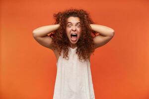 Teenage girl, unhappy looking woman with curly ginger hair. Wearing white off-shoulder blouse. Touching her head and screams, horrified. Watching at the camera, isolated over orange background photo