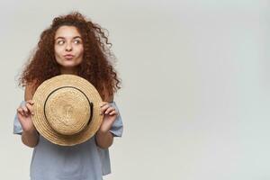 Nice looking woman, beautiful girl with curly ginger hair. Wearing striped off-shoulders blouse and holding a hat. Making a face. Watching to the right at copy space, isolated over white background photo