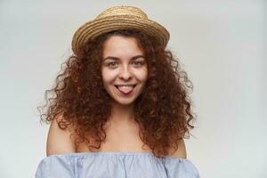 Portrait of attractive, adult redhead girl with curly hair. Wearing striped off-shoulders blouse and hat. Showing tongue. Watching at the camera, closeup, isolated over white background photo