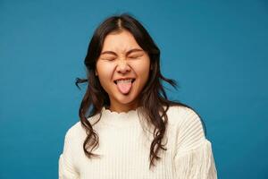 Good looking young pretty long haired woman with natural makeup keeping her eyes closed while making funny faces, showing her tongue while standing over blue background photo