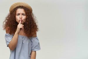 Teenage girl, woman with curly ginger hair. Wearing striped off-shoulders blouse and hat. Showing silence sign, frown. Watching at the camera, isolated over white background. Copy space on the right photo