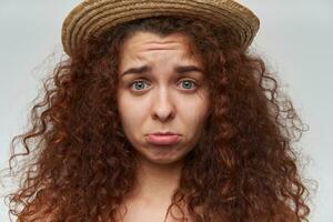 Sad looking woman, beautiful girl with curly ginger hair. Wearing a hat. Pout her lip and looking upset. Emotional concept. Watching at the camera, closeup, isolated over white background photo