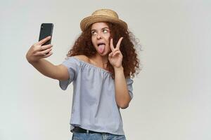 Silly looking woman, girl with curly ginger hair. Wearing striped off-shoulders blouse and hat. Taking a selfie on a smartphone, show peace sign and tongue. Stand isolated over white background photo