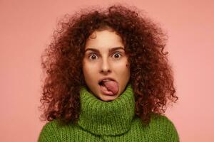 Portrait of attractive, adult girl with ginger curly hair. Wearing green turtleneck sweater and showing her tongue. Watching at the camera isolated, closeup over pastel pink background photo