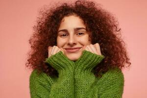 Young lady, pretty woman with ginger curly hair. Wearing green turtleneck sweater, feels warm and touching her cheeks, smiling. Watching at the camera isolated, closeup over pastel pink background photo