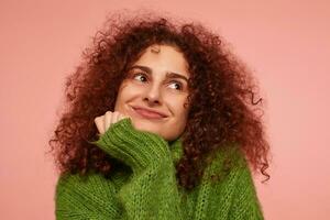 Portrait of attractive girl with ginger curly hair. Wearing green turtleneck sweater and dreaming, rests the chin. Watching to the right at copy space, isolated, closeup over pastel pink background photo