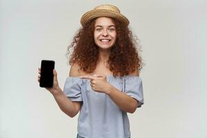 Nice looking woman, girl with curly ginger hair. Wearing striped off-shoulders blouse and hat. Holding and pointing at smartphone, copy space. Watching at the camera, isolated over white background photo