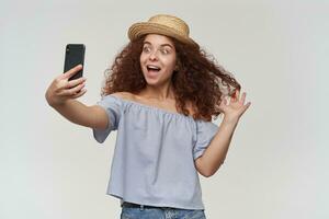 Portrait of attractive, redhead girl with curly hair. Wearing striped off-shoulders blouse and hat. Taking a selfie on a smartphone, play with hair and smile. Stand isolated over white background photo