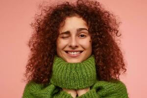 Young lady, pretty woman with ginger curly hair. Wearing green turtleneck sweater and put hands under it. Watching at the camera and wink, isolated, closeup over pastel pink background photo