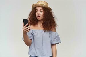 Teenage girl, happy looking woman with curly ginger hair. Wearing striped off-shoulders blouse and hat. Holding and watching at her smartphone, unhappy face. Stand isolated over white background photo