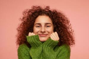 Teenage girl, happy looking redhead woman with curly hair. Wearing green turtleneck sweater and touching her cheeks, smiling. Watching at the camera isolated, closeup over pastel pink background photo