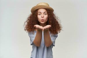 Nice looking woman, beautiful girl with curly ginger hair. Wearing striped off-shoulders blouse and hat. Sending air kiss to you. Watching at the camera, isolated over white background photo