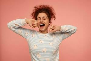 Portrait of attractive, adult girl with ginger curly hair. Wearing striped sweater with bunnies and yawns with closed eyes, stretching. Feels sleepy. Stand isolated over pastel pink background photo