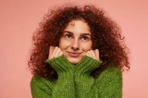 Portrait of attractive, adult girl with ginger curly hair. Wearing green turtleneck sweater and feels warm, touching her cheeks. Watching up isolated, closeup over pastel pink background photo