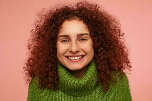 Teenage girl, happy looking redhead woman with curly hair. Wearing green turtleneck sweater and have a foxy smile. Watching at the camera isolated, closeup over pastel pink background photo