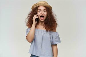 Portrait of attractive, adult redhead girl with curly hair. Wearing striped off-shoulders blouse and hat. Talk on a phone, hear a great news. Watching at the camera, isolated over white background photo