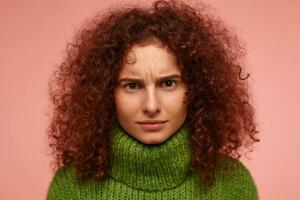 Portrait of attractive, adult girl with ginger curly hair. Unsatisfied looking. Wearing green turtleneck sweater and frowns watching at the camera isolated, closeup over pastel pink background photo