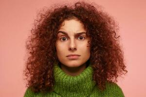 Teenage girl, serious looking redhead woman with curly hair. Wearing green turtleneck sweater and lifts up an eyebrow. Watching at the camera isolated, closeup over pastel pink background photo