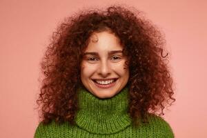 Teenage girl, happy looking redhead woman with curly hair. Wearing green turtleneck sweater and have a bright, warm smile. Watching at the camera isolated, closeup over pastel pink background photo