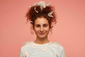 retrato de atractivo, adulto niña con jengibre Rizado cabello. vistiendo a rayas suéter con conejitos y cubierto con plumas, sonriente. acecho a el cámara aislado, de cerca terminado pastel rosado antecedentes foto