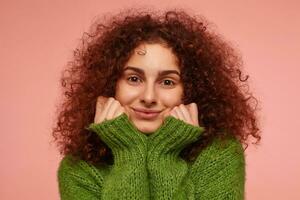 Teenage girl, happy looking redhead woman with curly hair. Wearing green turtleneck sweater and feels warm, touching her cheeks. Watching at the camera isolated, closeup over pastel pink background photo