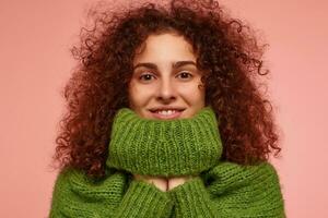 Teenage girl, happy looking redhead woman with curly hair. Wearing green turtleneck sweater and hide hands under turtleneck. Watching at the camera isolated, closeup over pastel pink background photo