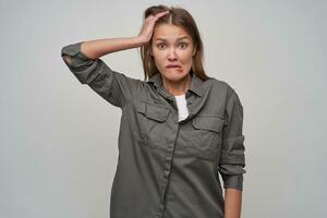 Teenage girl, scared looking woman with brown long hair. Wearing grey shirt and touching her head in shock, biting her lip. Watching at the camera isolated over grey background photo