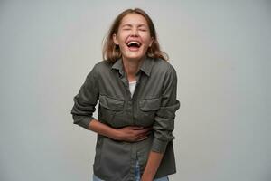 Portrait of attractive, adult girl with brown long hair, piercing. Wearing grey shirt and laughing laud with closed eyes, touching her belly. Stand isolated over grey background photo